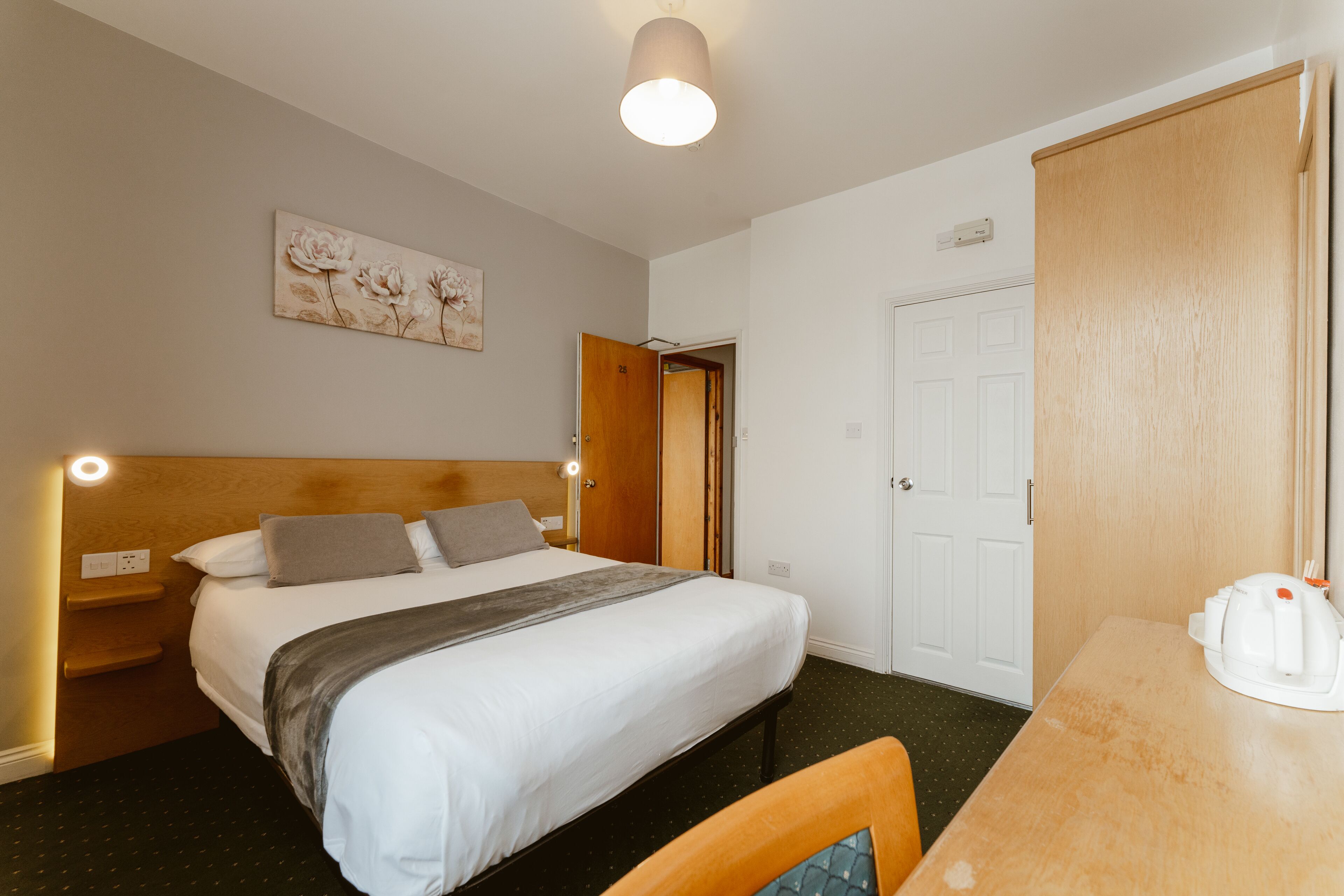 A hotel room with a bed, a desk, a chair, a door, and a wall-mounted light fixture.