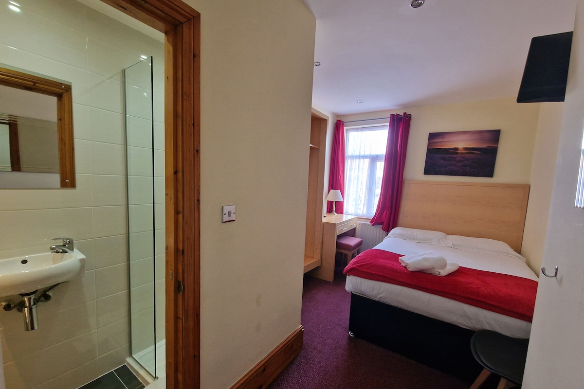 Habitación de hotel con ducha, cama con manta roja, escritorio con lámpara y un cuadro en la pared.