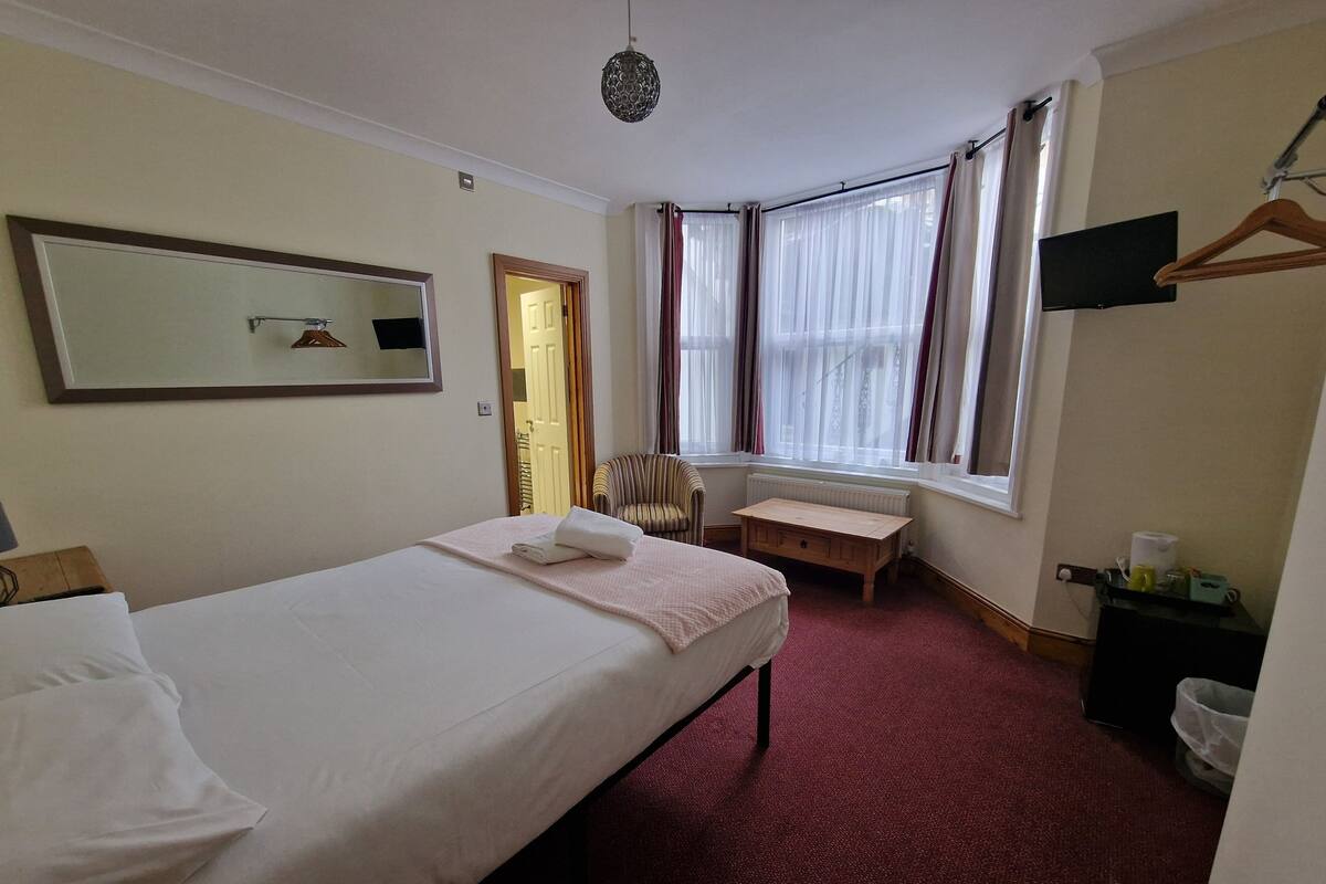 A hotel room with a bed, a chair, a small table, a mirror, and a window with curtains.