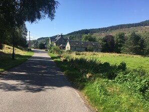 A rural road leading to a row of stone houses with a green roof, surrounded by lush greenery and hills. - The Tomich Hotel (Beauly)