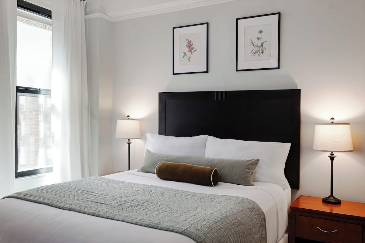 A neatly made bed with a white comforter, two pillows, and a brown throw. There are two bedside tables with lamps, and the room has a window with white curtains.