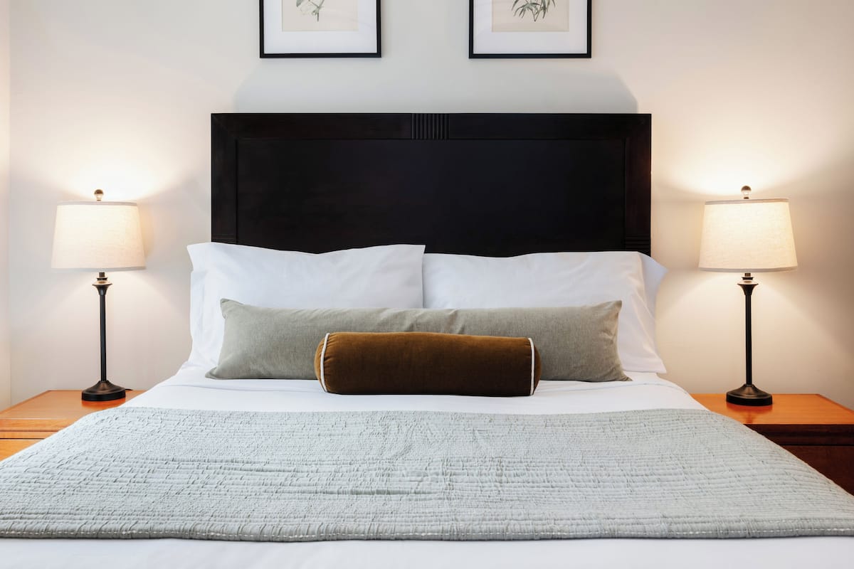 A neatly made bed with white and gray bedding, two pillows, and a brown accent pillow. There are two bedside lamps on wooden nightstands.