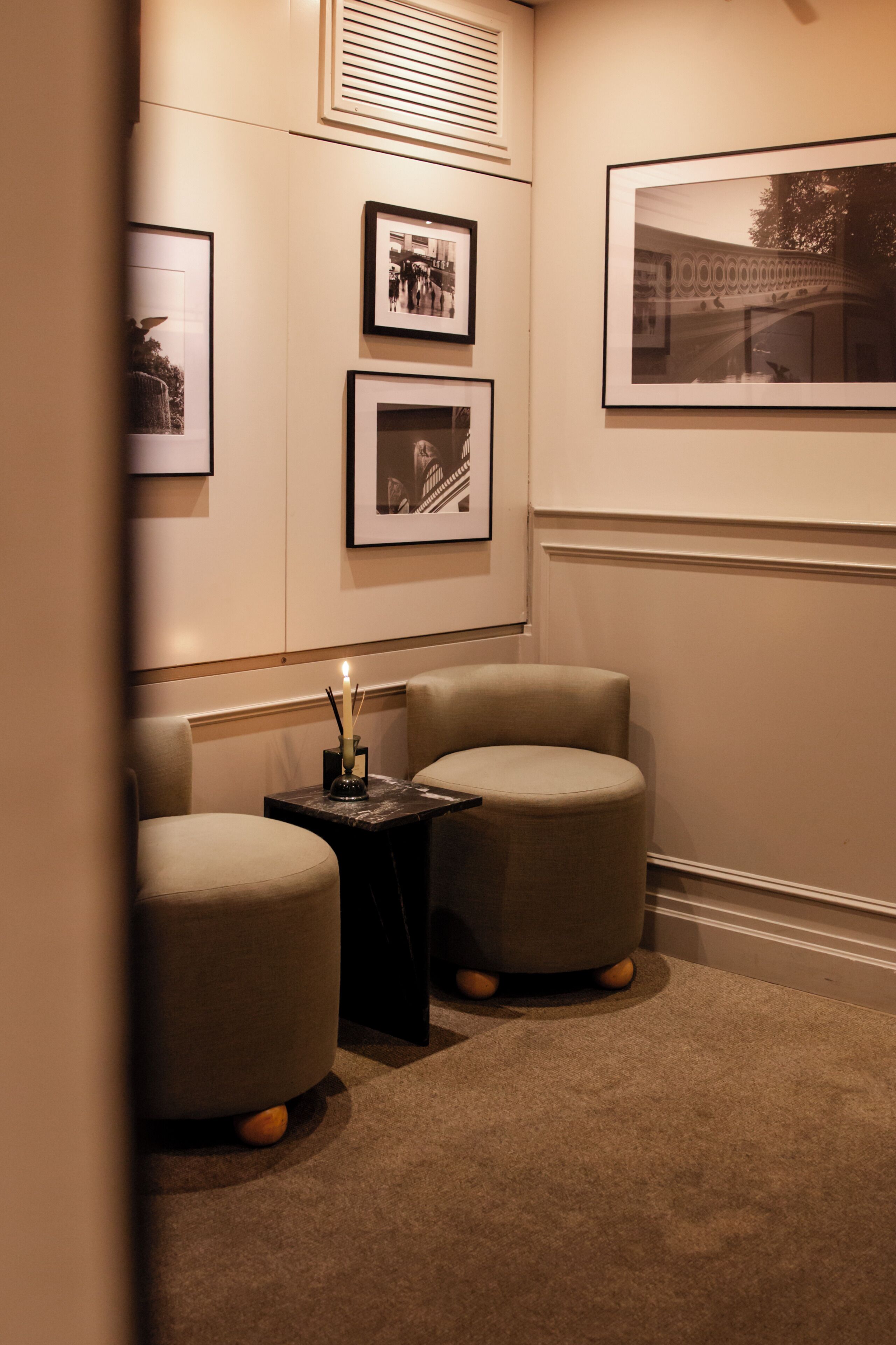 A corner room with two round ottomans, a small table with a candle, and framed pictures on the wall.
