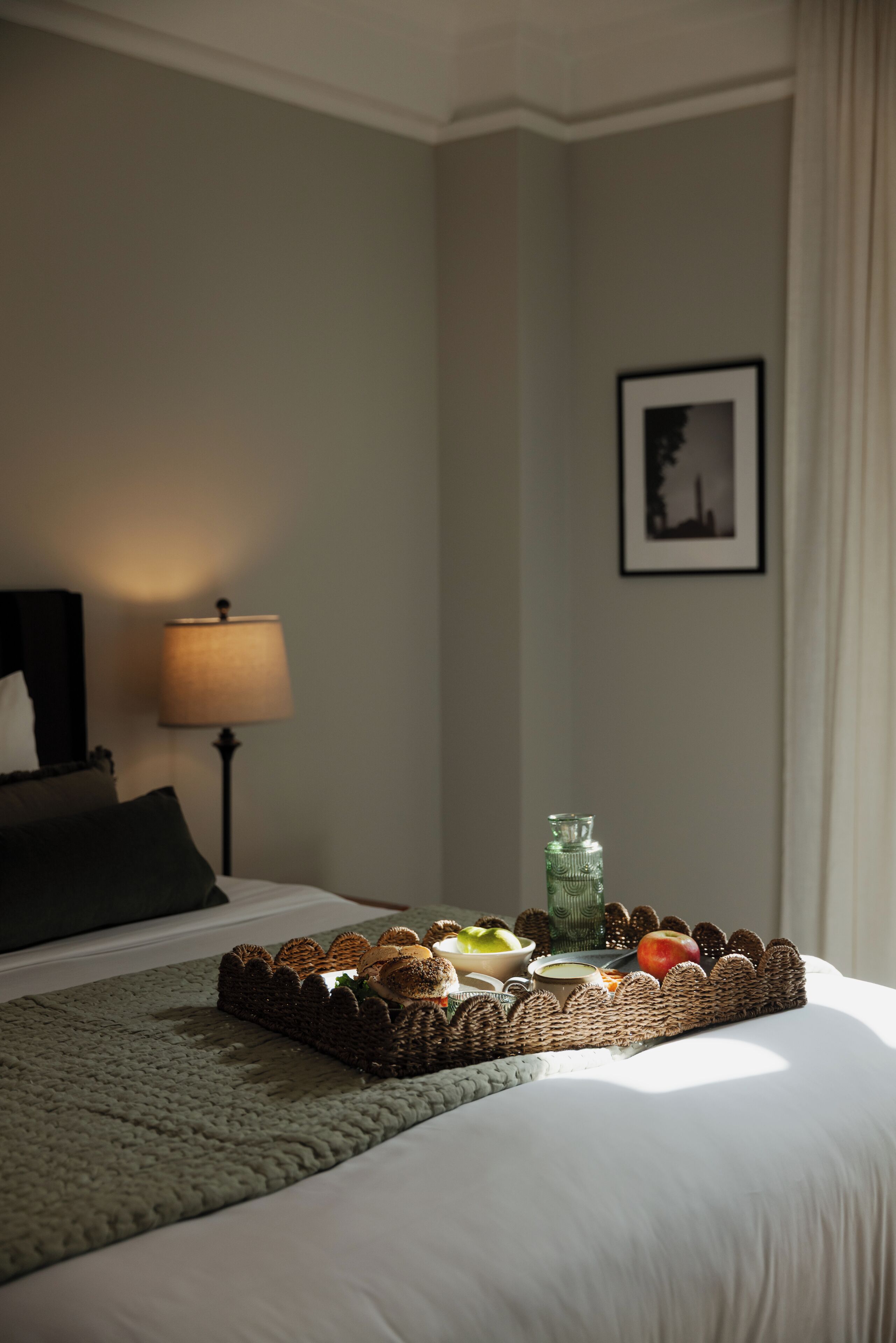 Un lit bien fait, avec un plateau contenant des articles pour le petit-déjeuner, une lampe et un tableau encadré accroché au mur.