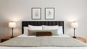 Ein ordentlich gemachtes Bett mit braun-weißem Kopfteil, zwei gerahmte botanische Drucke an der Wand und zwei Nachttische mit hölzernen Beinen, jeweils mit einer Nachttischlampe.