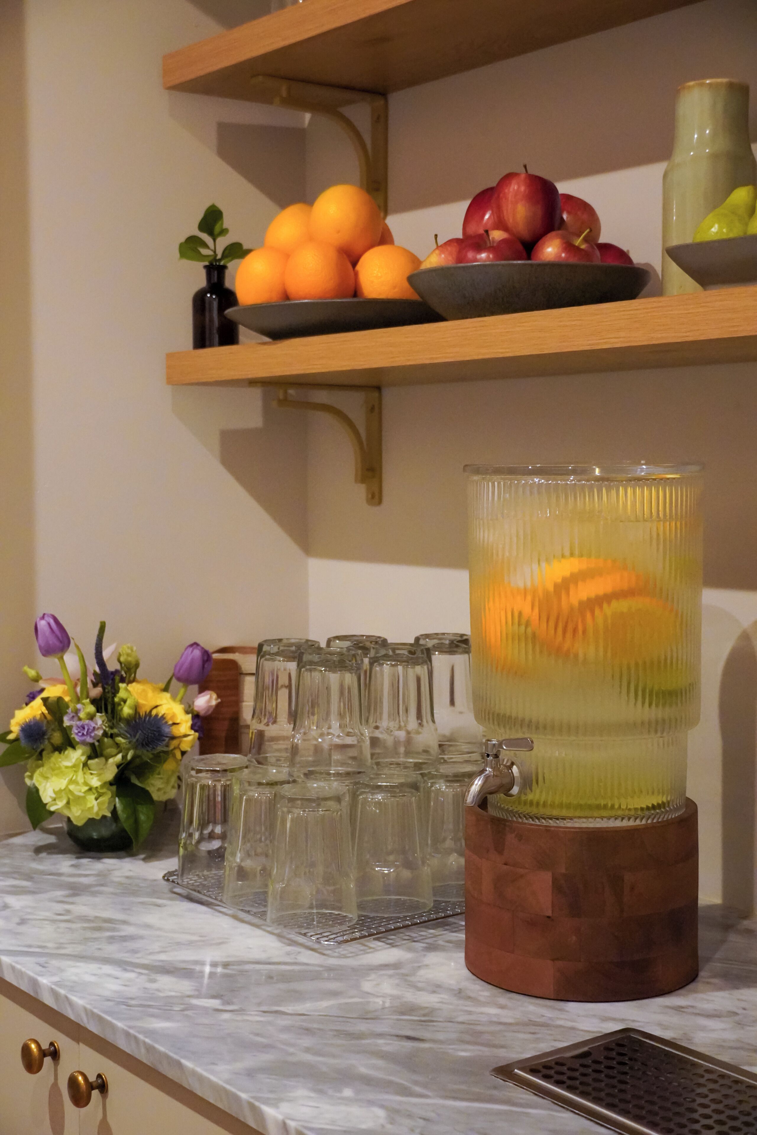 Eine Küchenarbeitsplatte mit einer Obstschale mit Orangen und Äpfeln, einer Blumenvase und einer Glas Karaffe.