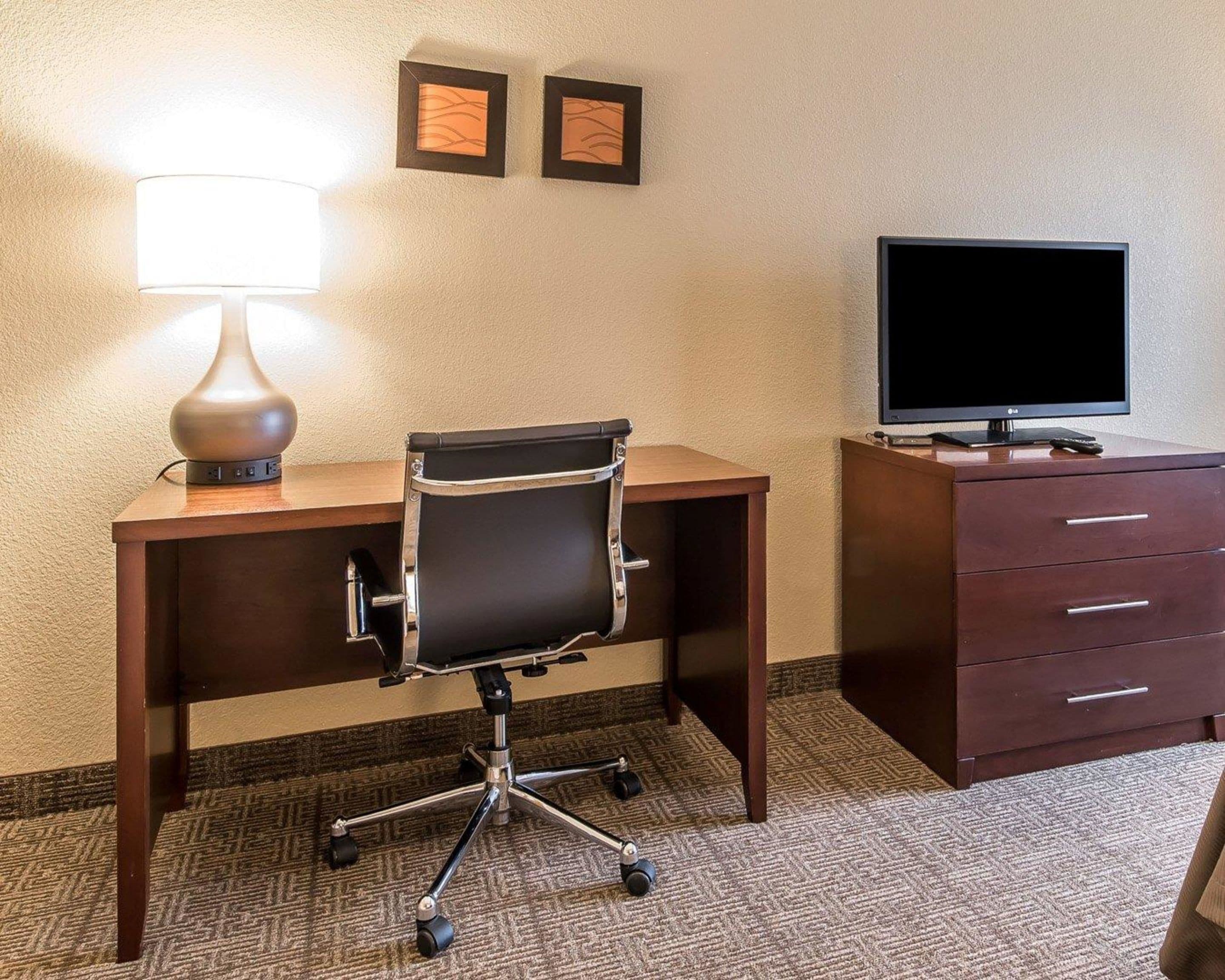 A hotel room with a desk, a chair, a lamp, a television, and a dresser.