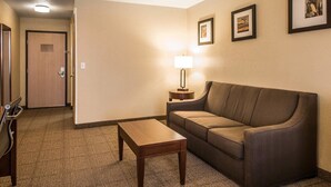 A hotel room with a brown leather sofa, a wooden coffee table, and a lamp on a side table. There are framed pictures on the wall and a door in the background.