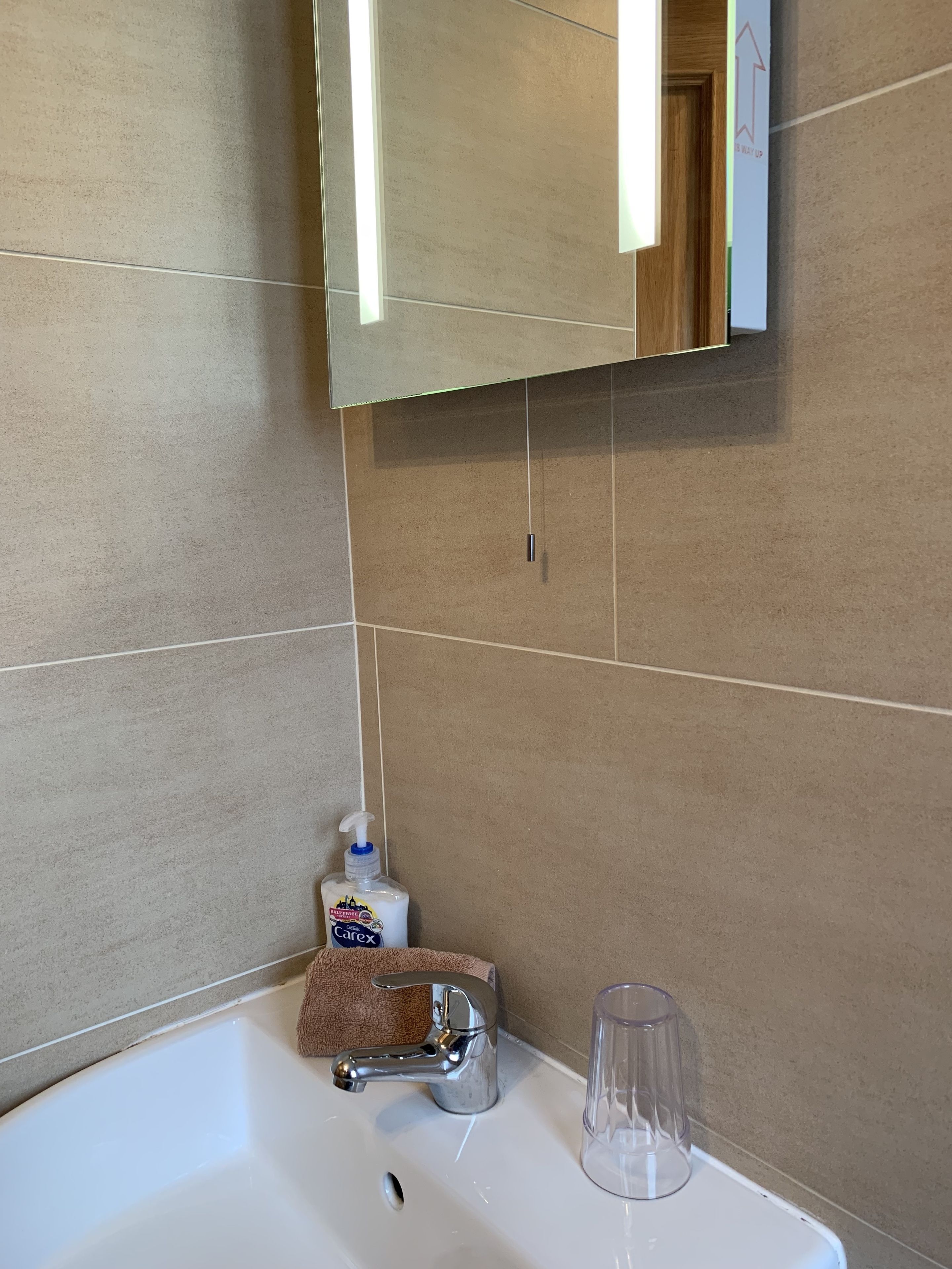 Un baño con un lavamanos blanco, un espejo, una botella de jabón para manos, una toalla y un vaso de vidrio.