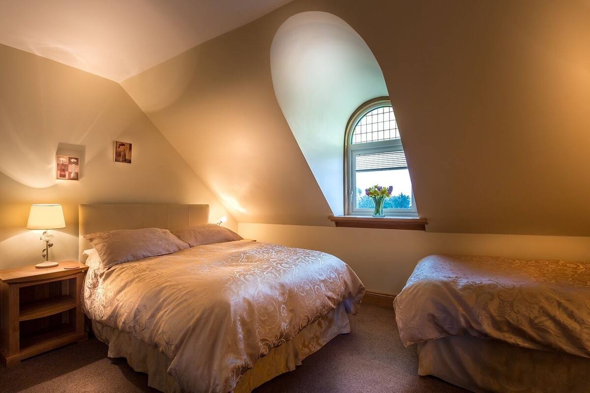 Ein Schlafzimmer mit zwei Betten, einem Nachttisch, einer Lampe und einem Fenster mit Blick ins Grüne.