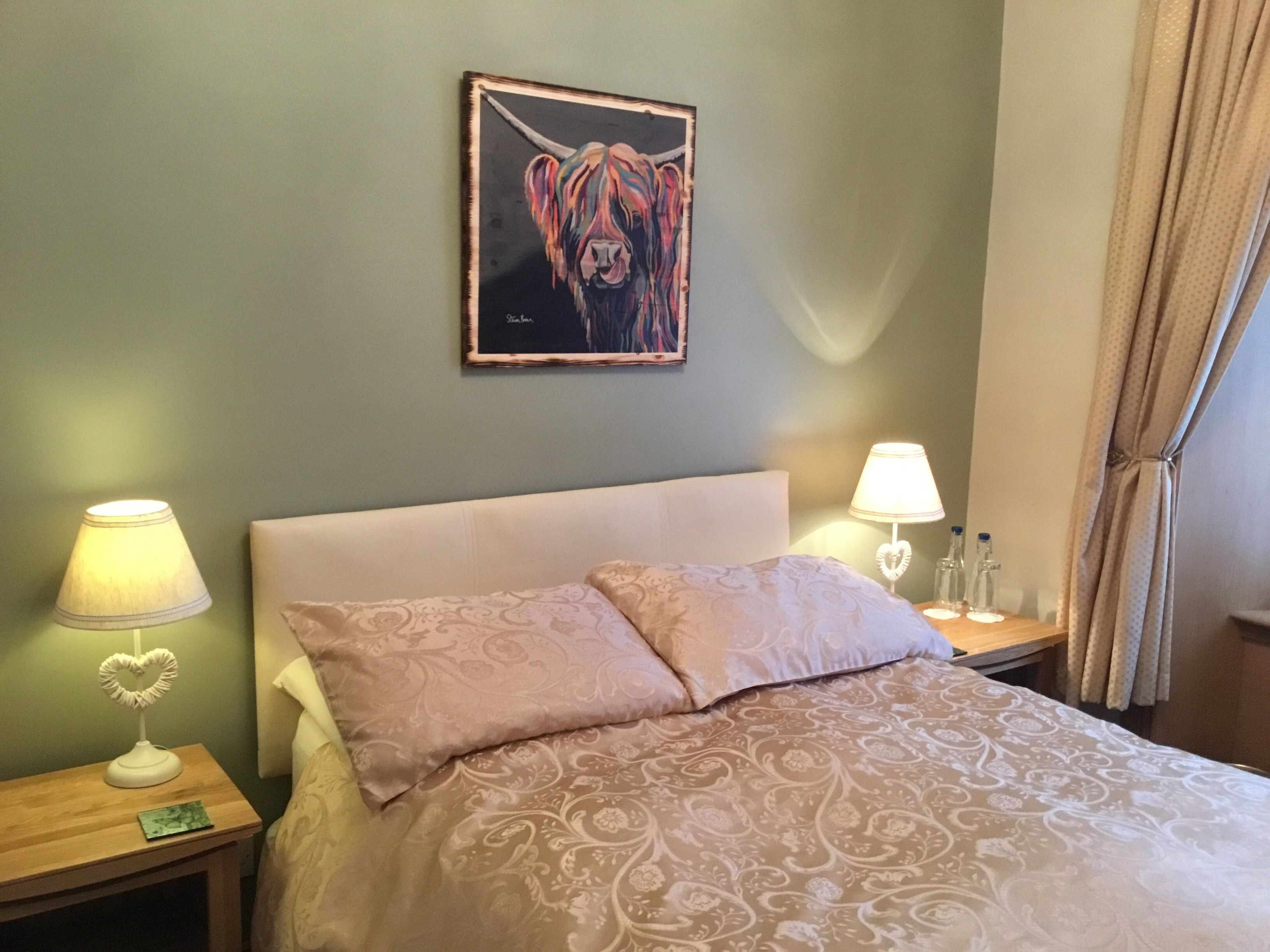 A hotel room with a bed, two bedside tables, a heart-shaped lamp, a painting of a cow, and a curtain.
