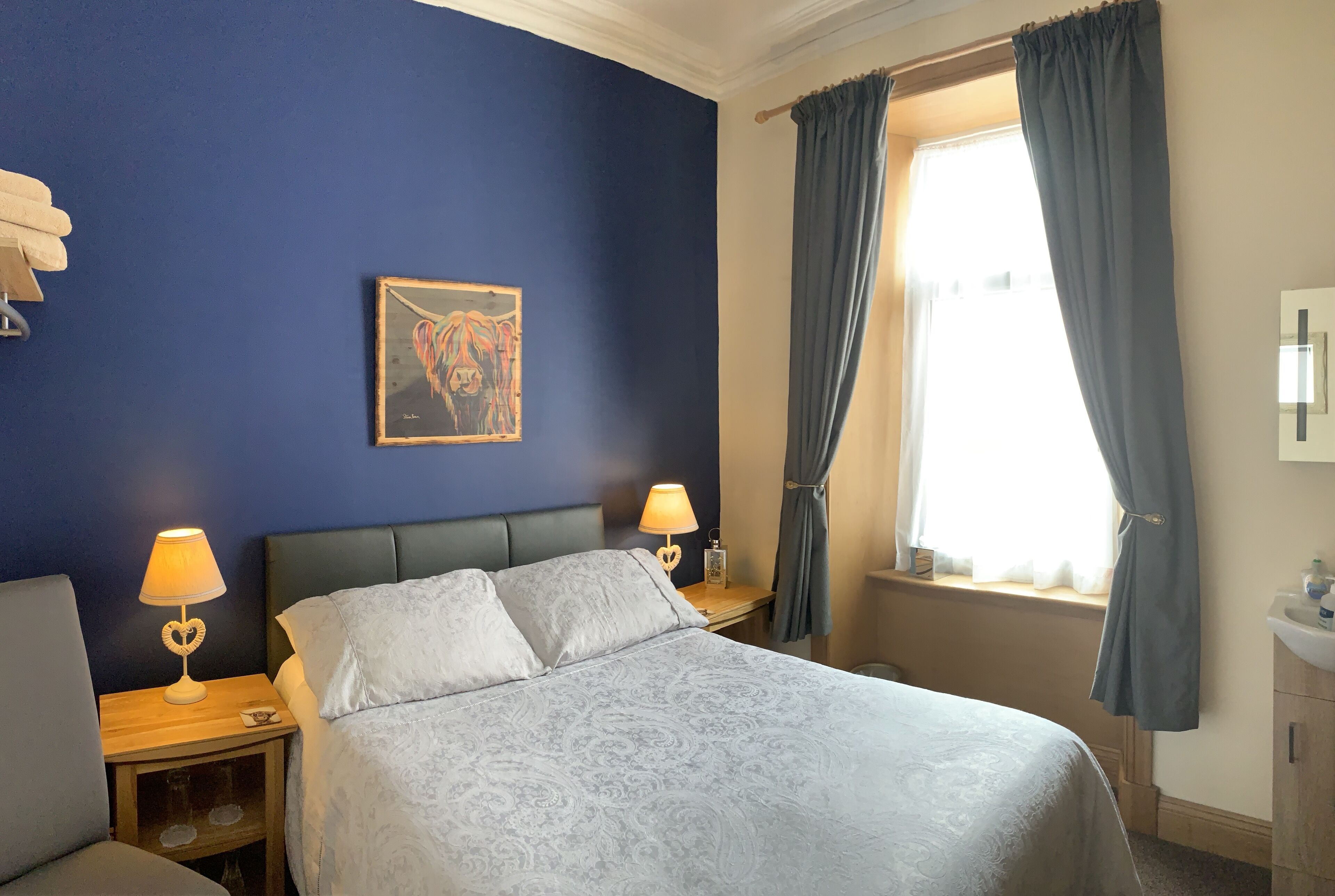Quarto com cama, duas mesinhas de cabeceira com luminárias, janela com cortinas, um quadro na parede e pia no canto.