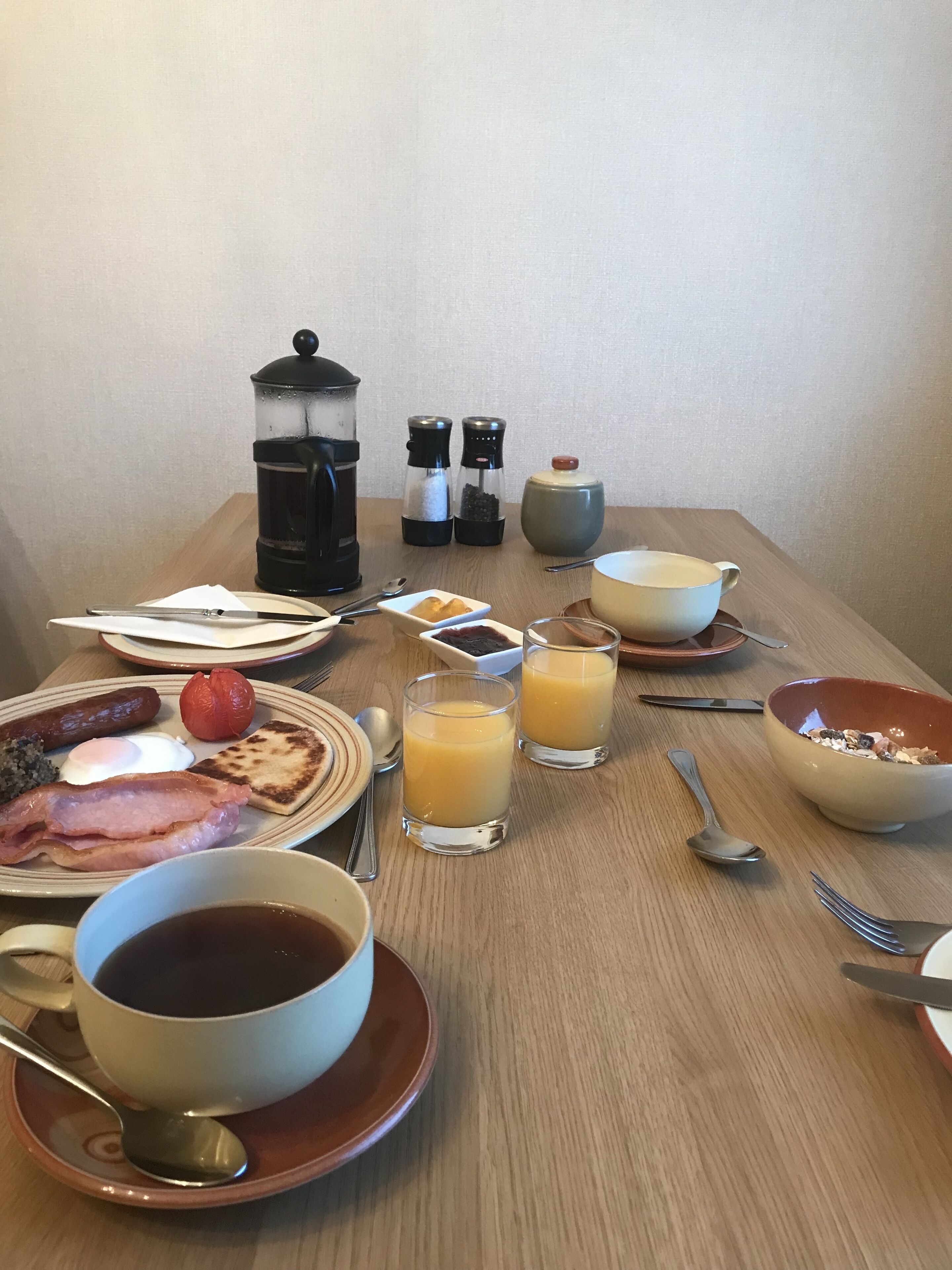 A breakfast table with plates of eggs, bacon, toast, and fruit; two glasses of orange juice; a cup of coffee; and a coffee pot.