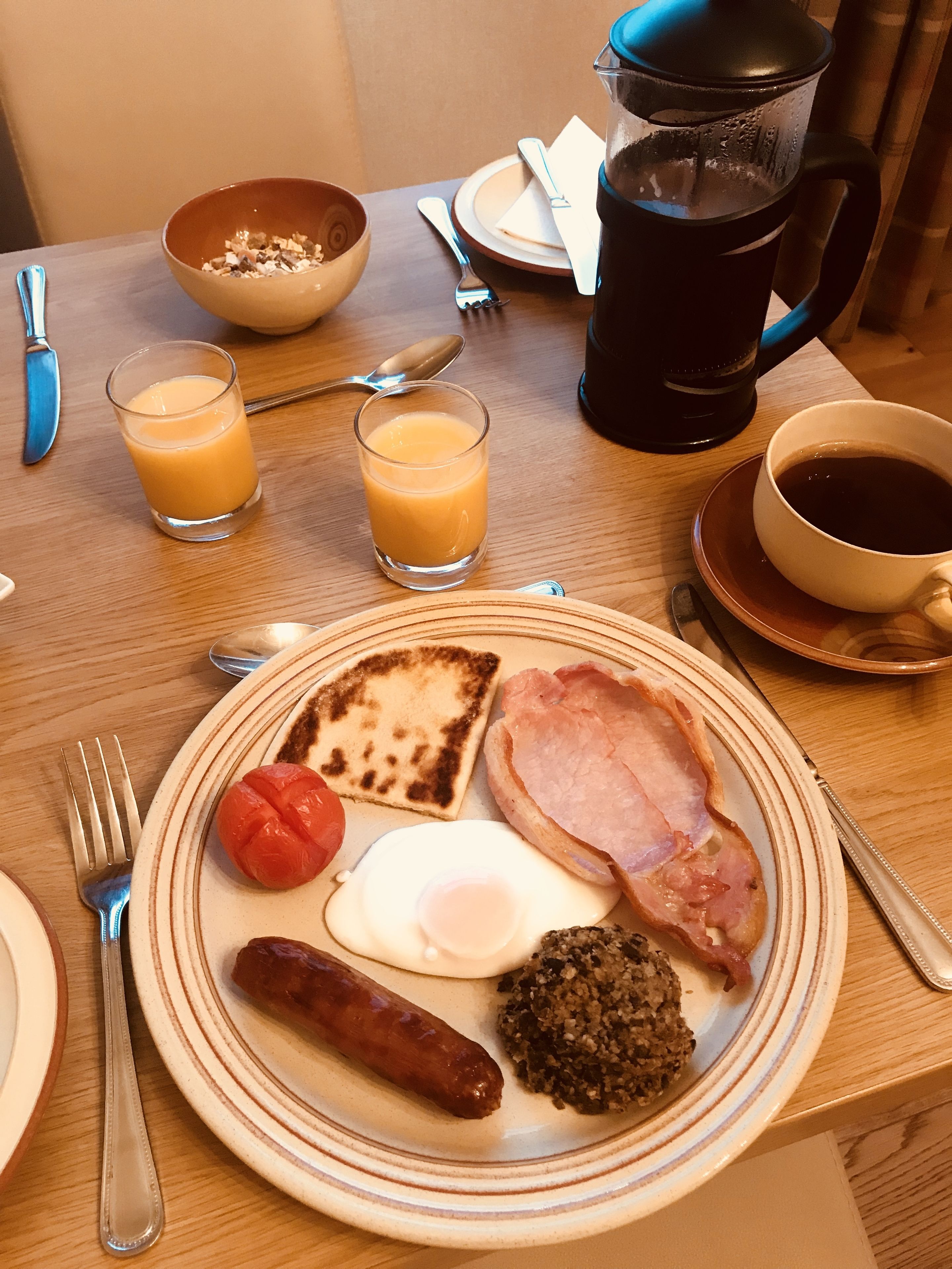 A breakfast table with toast, eggs, sausage, tomatoes, and hash browns. There are also two glasses of orange juice and a cup of coffee.