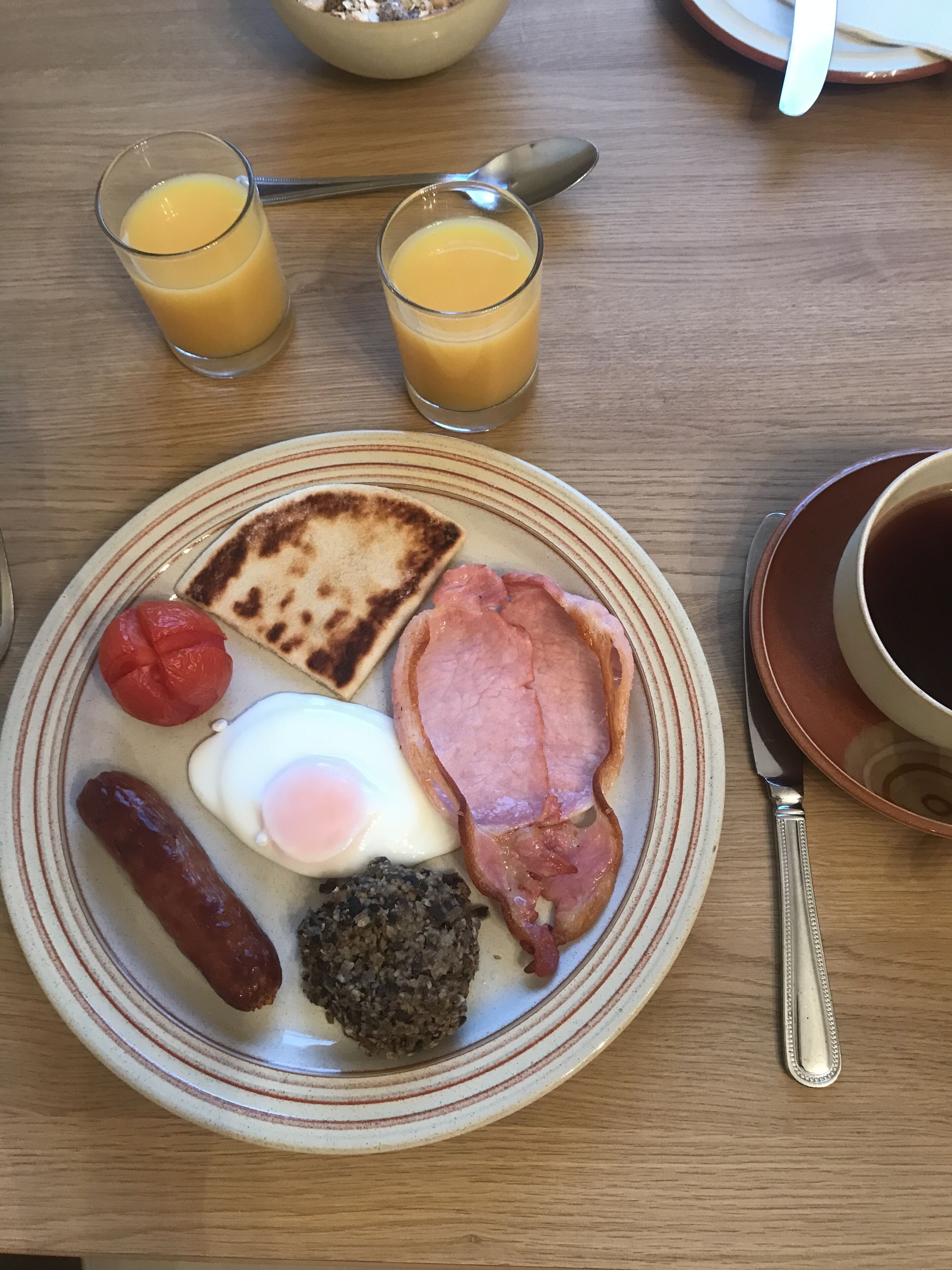 Un piatto per la colazione con pane tostato, prosciutto, uova, salsiccia e un frutto rosso su un piatto di ceramica. Due bicchieri di succo d'arancia sono sul tavolo.