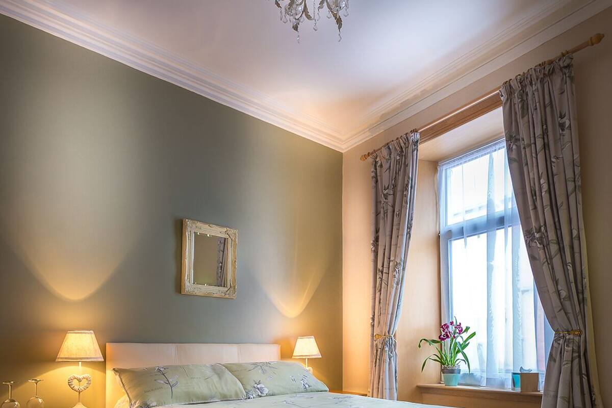 Ein Schlafzimmer mit einem großen Bett, zwei Nachttischen mit Lampen, einem Kronleuchter, einem Fenster mit Vorhängen und einem gerahmten Bild an der Wand.