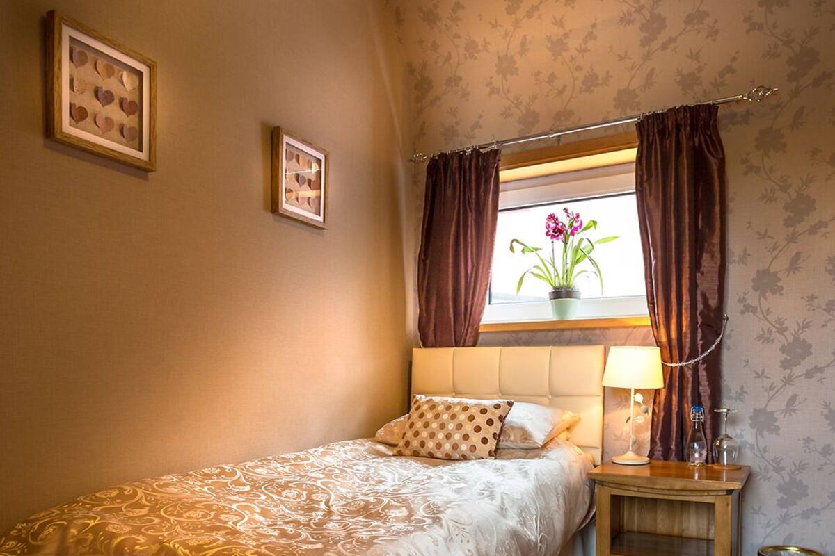 Ein Schlafzimmer mit Bett, Nachttisch, Lampe, Fenster mit Vorhängen und Wand mit floralem Tapetenmuster.