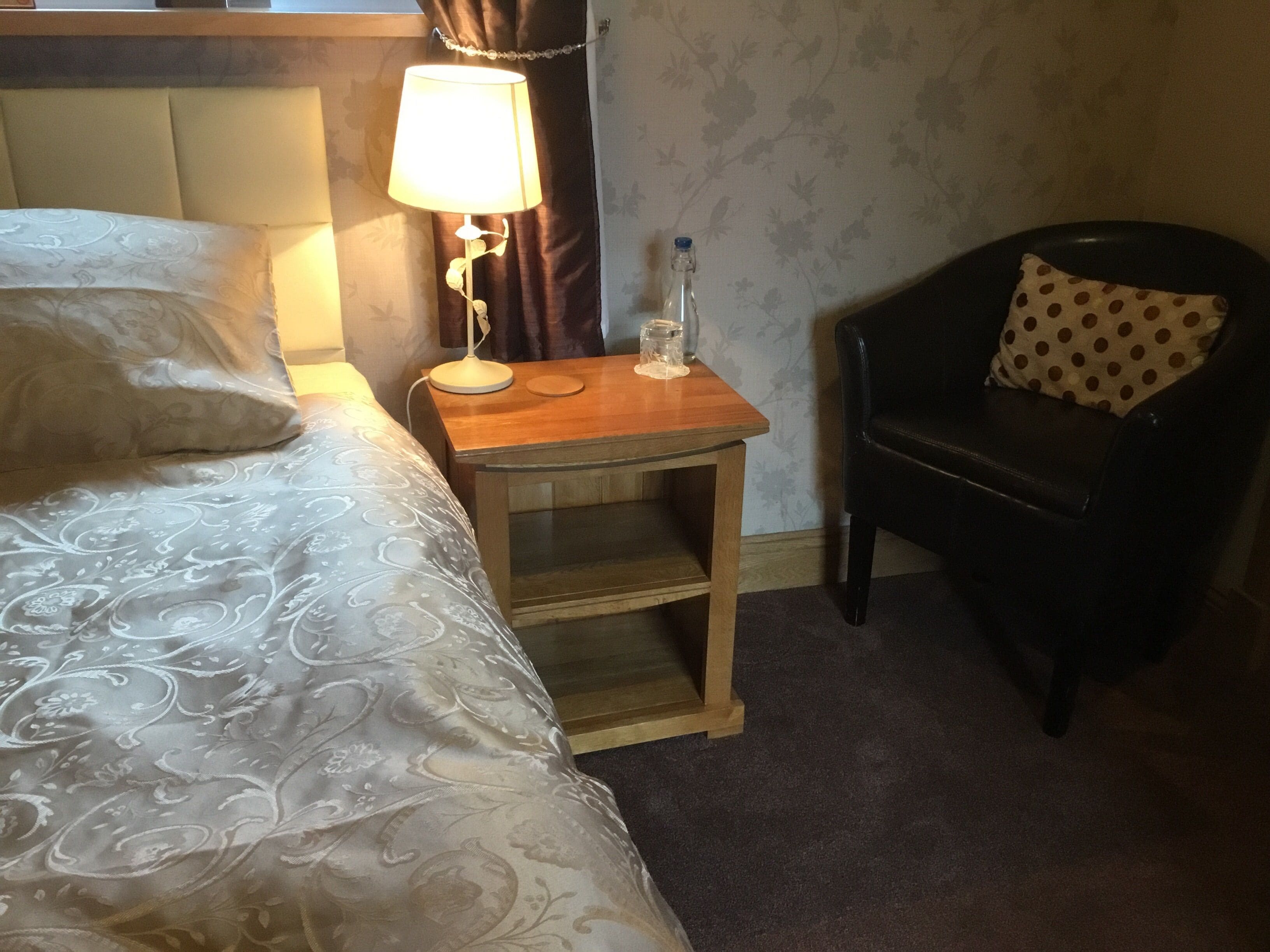 Una habitación de hotel con cama, una mesita de noche con lámpara, una botella y una silla con un cojín estampado de lunares.