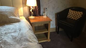 Una habitación de hotel con cama, una mesita de noche con lámpara, una botella y una silla con un cojín estampado de lunares.