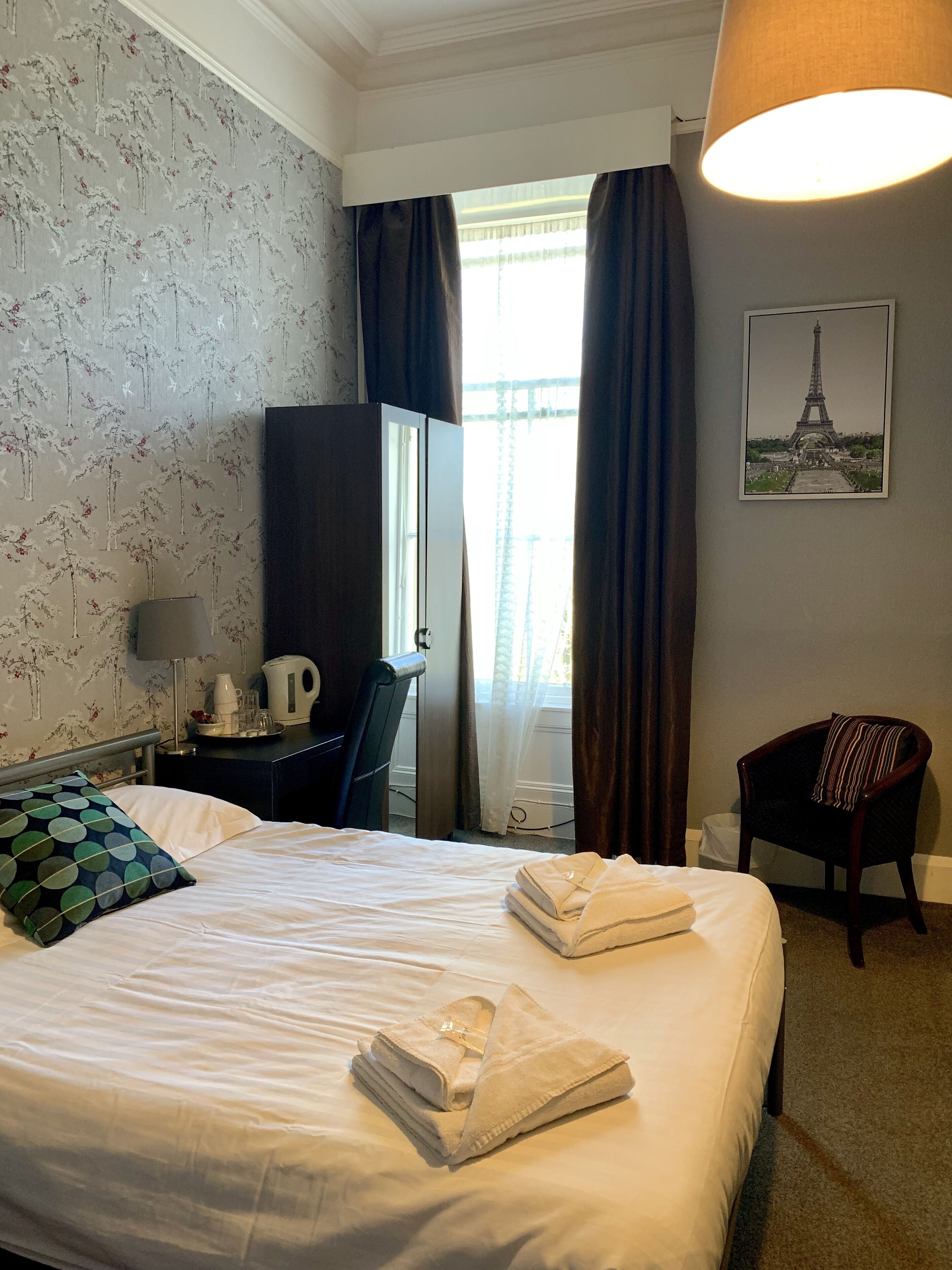 A hotel room with a bed, a desk, a chair, a lamp, and a window with curtains.