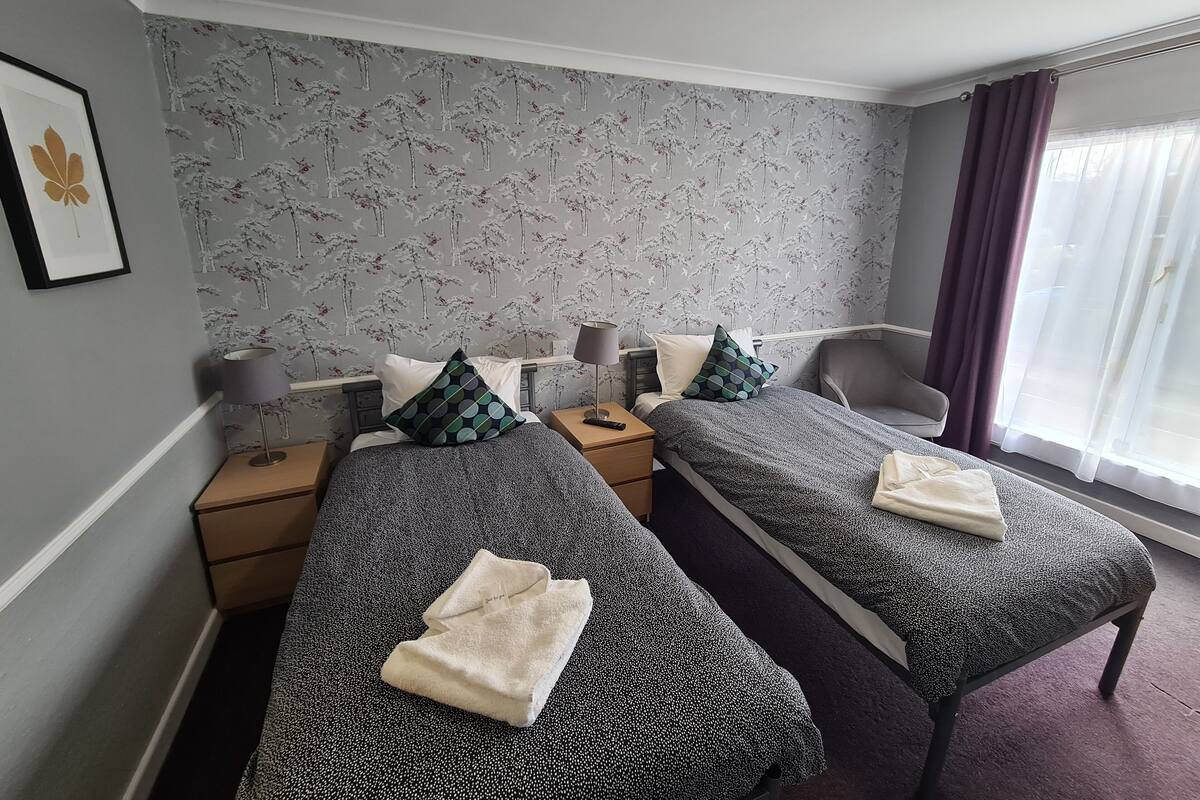 A hotel room with two single beds, a desk, a chair, and a window with curtains.