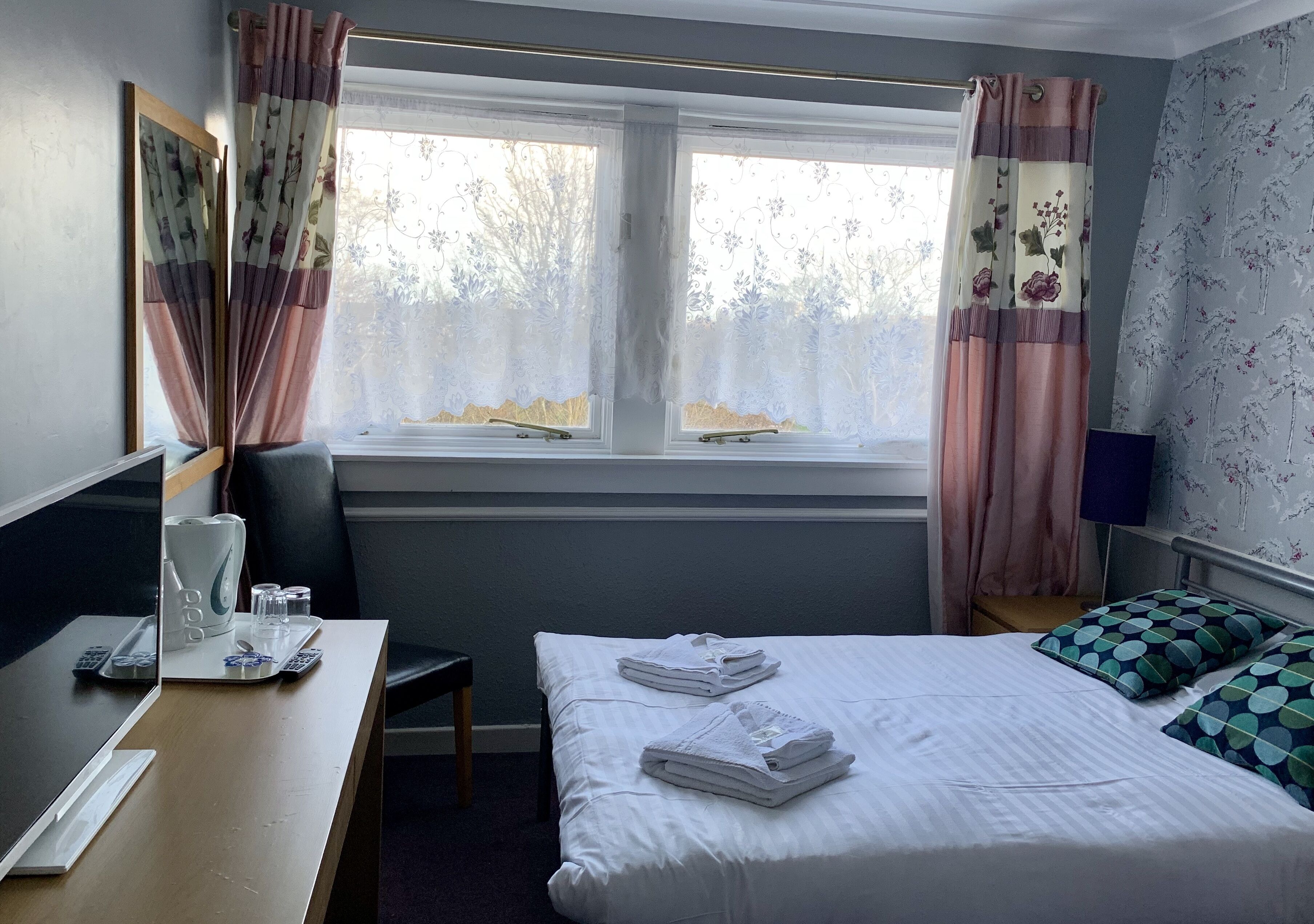 A hotel room with a bed, a desk with a kettle, glasses, and a tray, and a window with curtains.