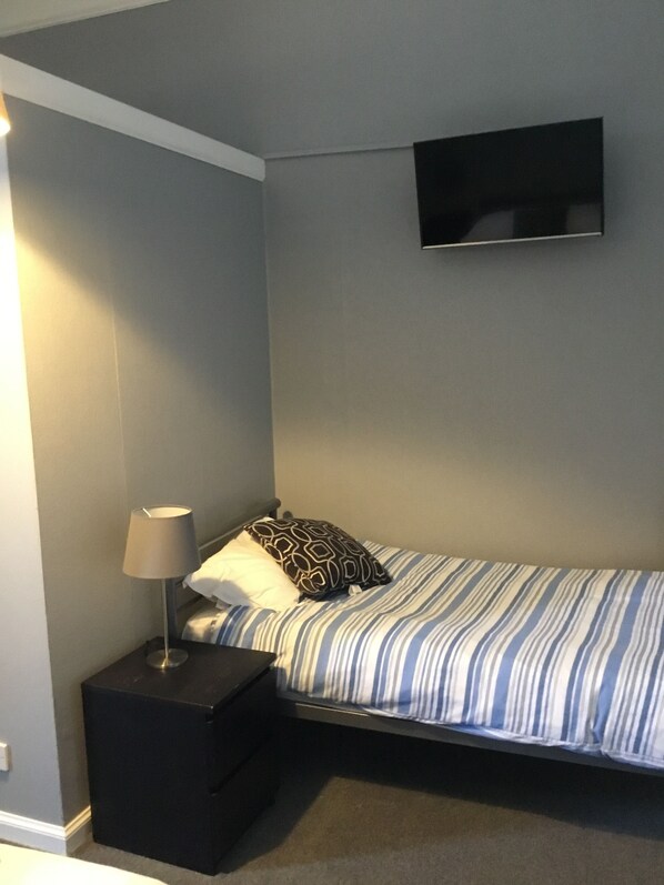 Una habitación pequeña con una cama individual, una colcha a rayas, una mesita de noche negra con una lámpara y un televisor de pantalla plana montado en la pared.