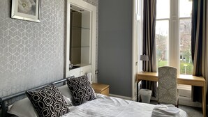 A hotel room with a bed, a bedside table, a chair, a desk, a window with curtains, and a wall with a geometric pattern. - Northumberland Hotel (Edinburgh)