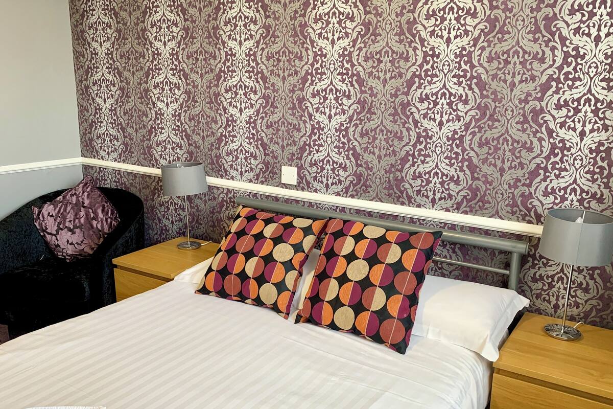 A neatly made bed with patterned pillows, a bedside lamp, and a chair with a cushion.