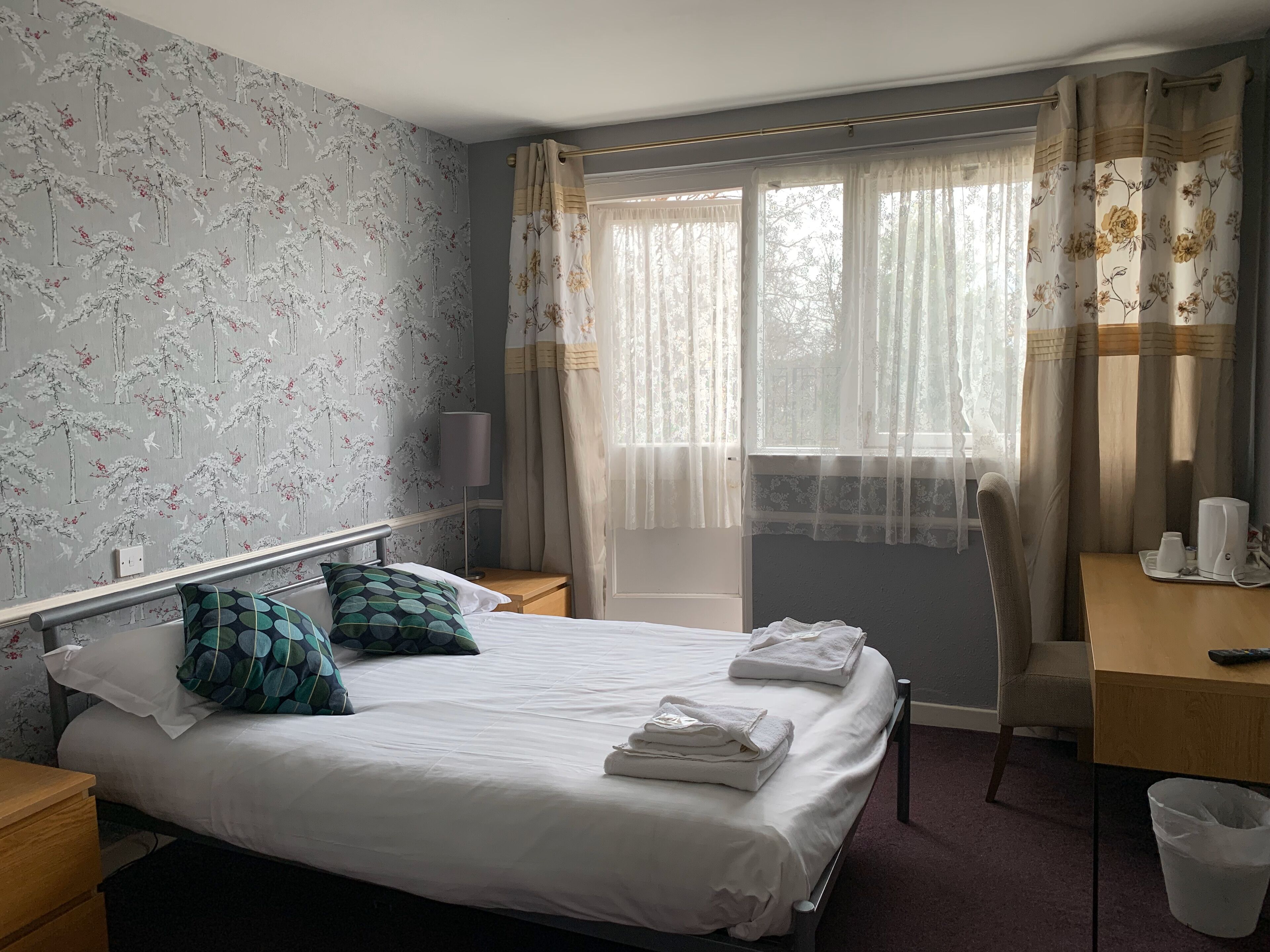 A hotel room with a bed, a desk, a chair, a lamp, and a window with curtains.