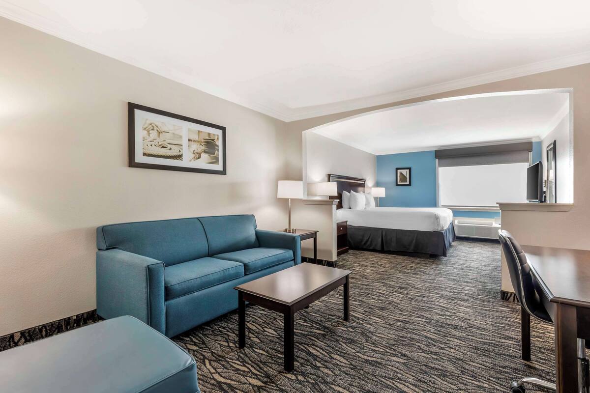 A hotel room with a blue sofa, a small table, a bed with a white comforter, a desk with a chair, and a large mirror.