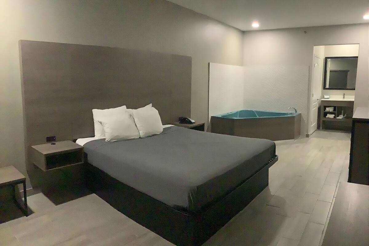 Habitación de hotel moderna con una cama grande, mesitas de noche, un escritorio, una silla, una mesita pequeña, un espejo y un baño con lavabo y ducha.