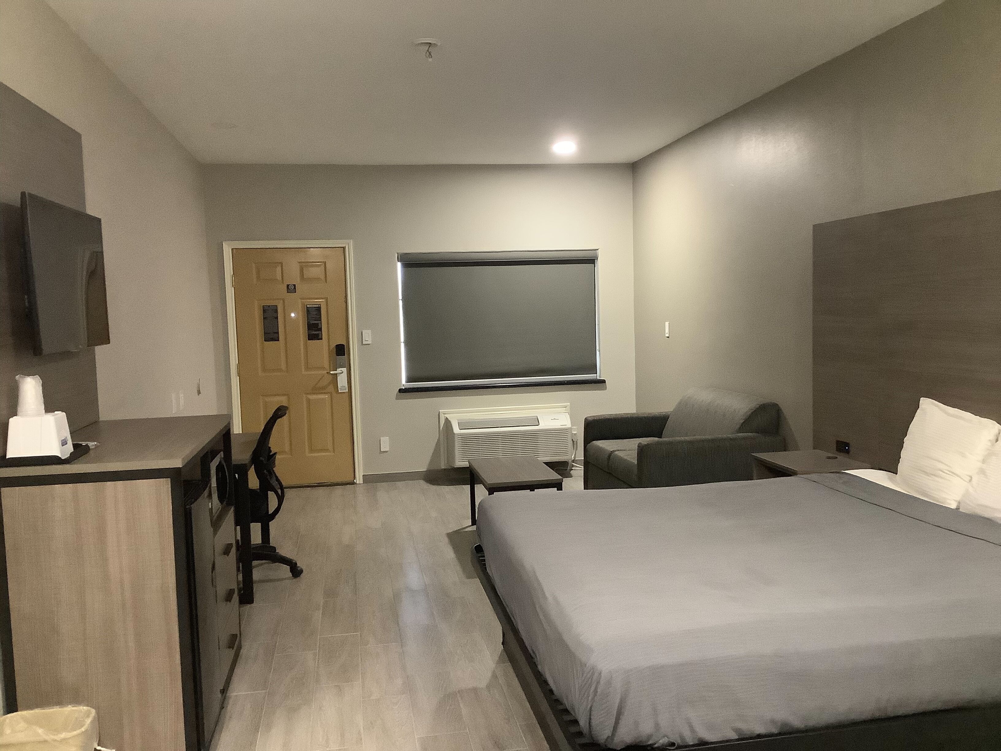A hotel room with a bed, a desk, a chair, a TV mounted on the wall, and a door.
