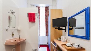 Une chambre d’hôtel compacte comprenant un lavabo, un miroir, une serviette rouge, une télévision et un petit bureau avec une bouilloire.