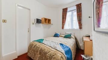 A bedroom with a bed, a window with curtains, a bedside table, and a trash bin.