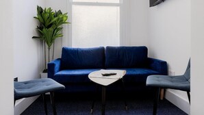 Une pièce avec un canapé bleu, deux fauteuils gris, une petite table blanche, une plante en pot et un téléviseur fixé au mur.