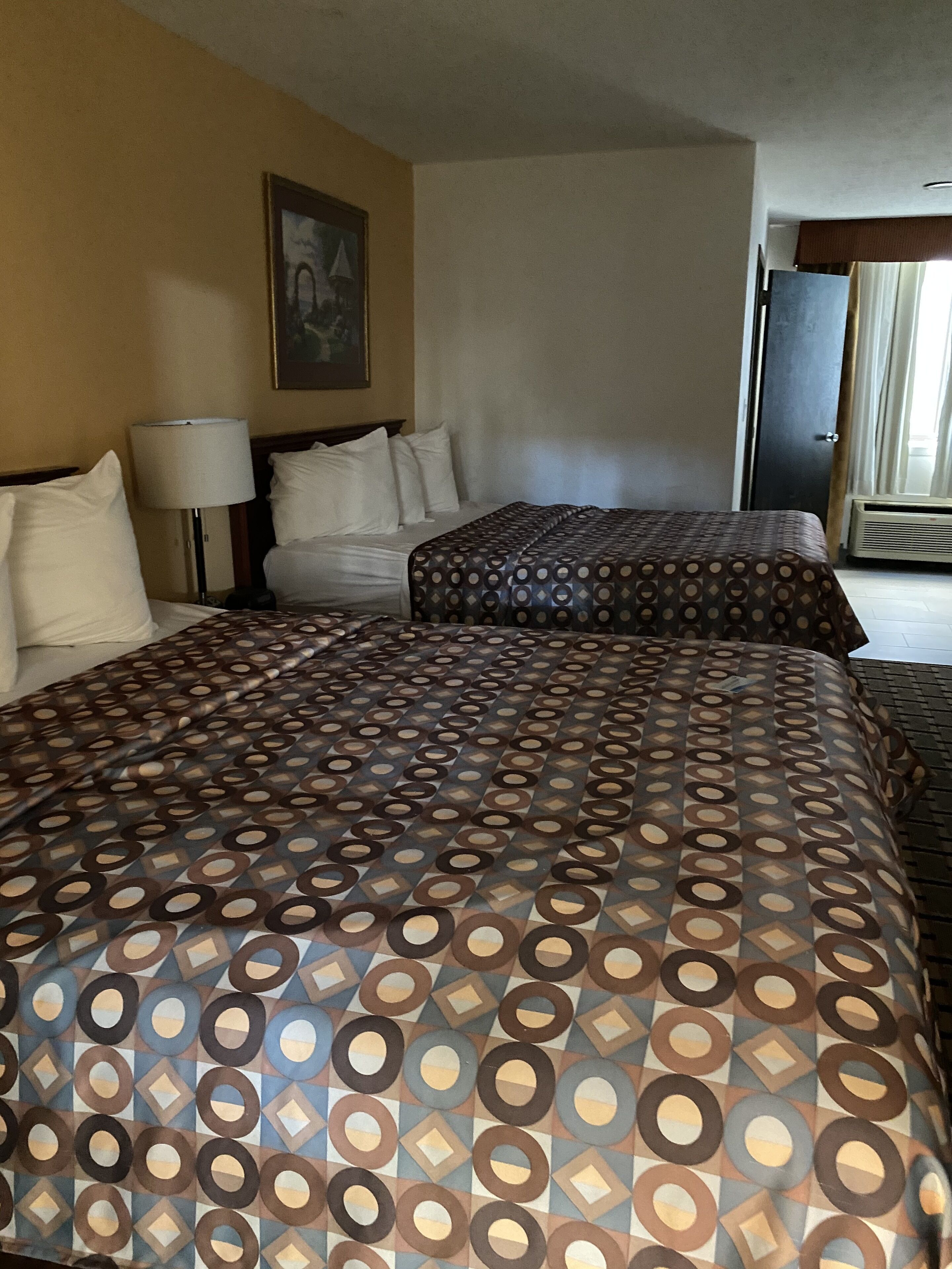 A hotel room with two beds, a nightstand, a lamp, and a door.