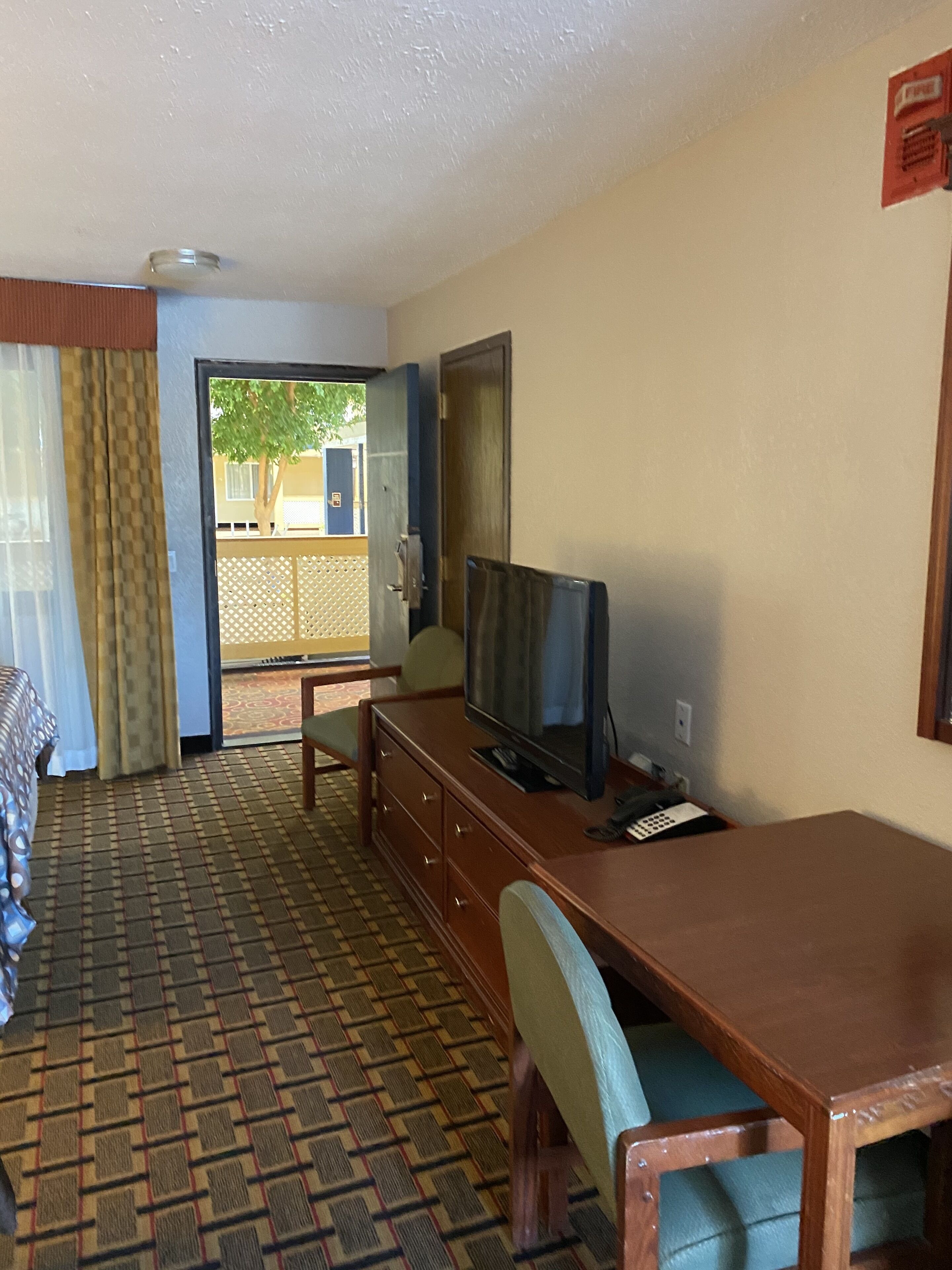 Habitación de hotel con un escritorio de madera, televisión, silla, alfombra estampada, ventana con cortinas y puerta que da al exterior.