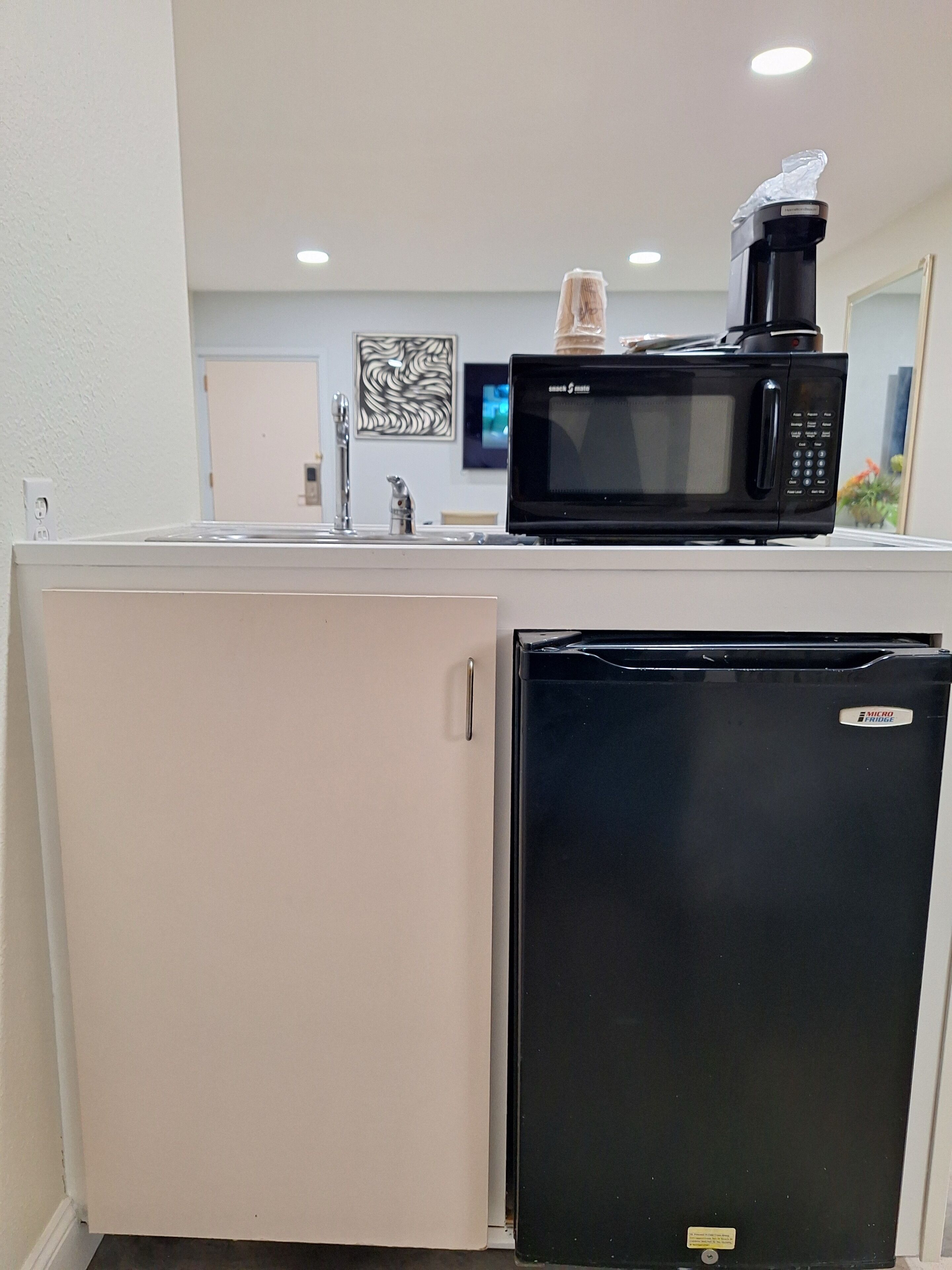 Una kitchenette compacta con un microondas negro, un refrigerador pequeño y una cafetera sobre la encimera.