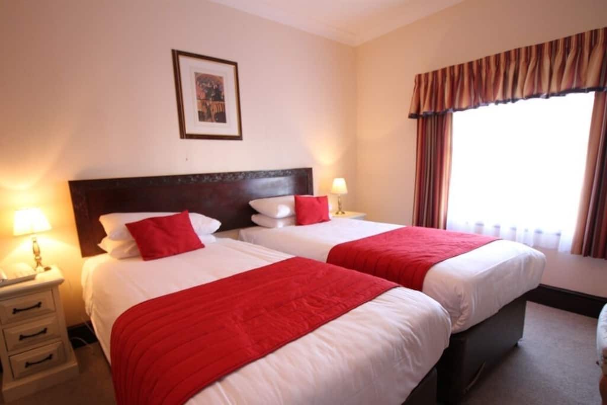 A hotel room with two beds, each with white bedding and red accents, a wooden headboard, bedside lamps, a framed picture on the wall, and a window with curtains.