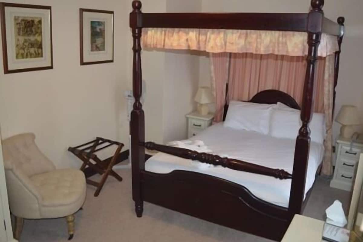 A four-poster bed with a canopy, a bedside table, a chair, and two framed pictures on the wall.