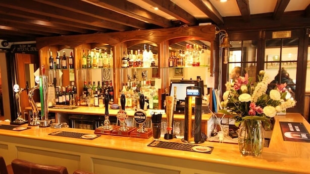 A bar area with wooden beams, a variety of liquor bottles, a cash register, and a vase with flowers. - Pilgrims Hotel (Canterbury)