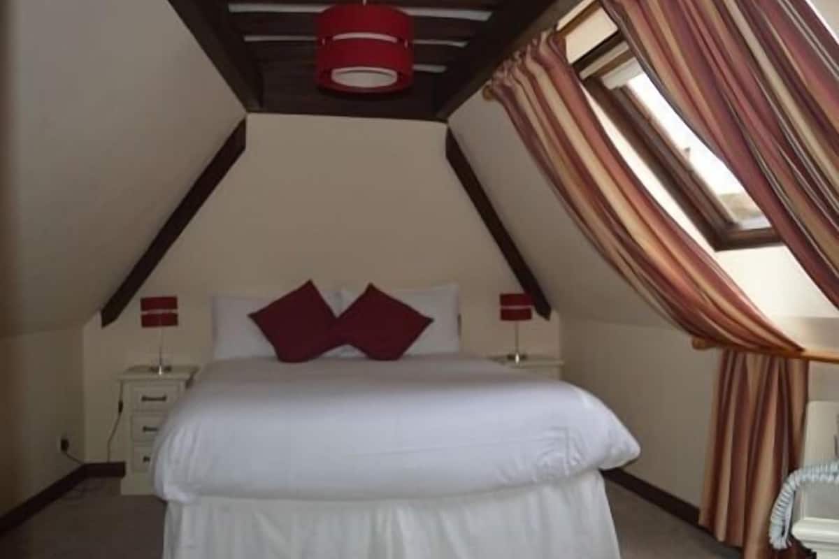 A bedroom with a sloped ceiling, a bed with white bedding, two red lamps, and a window with curtains.