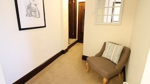 A hallway with a framed drawing, a window with blinds, and a brown armchair with a striped pillow. - Pilgrims Hotel (Canterbury)