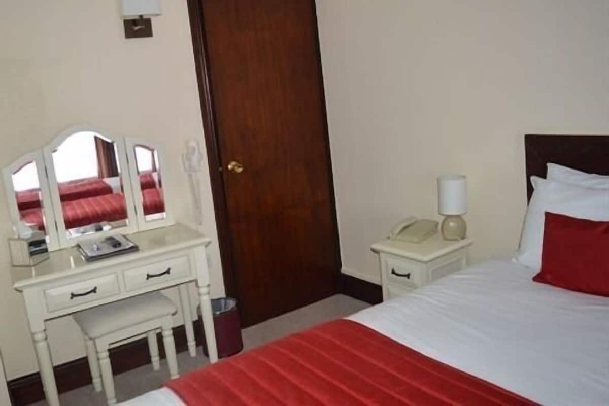 A hotel room with a bed, a bedside table with a lamp, a telephone, a mirror, and a small trash bin.