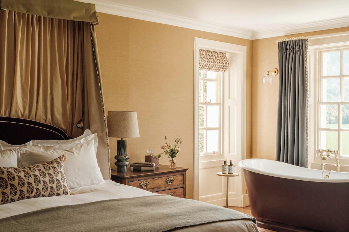 Een slaapkamer met een groot bed, een nachtkastje, een lamp, een vaas met bloemen, een raam met gordijnen en een bad.