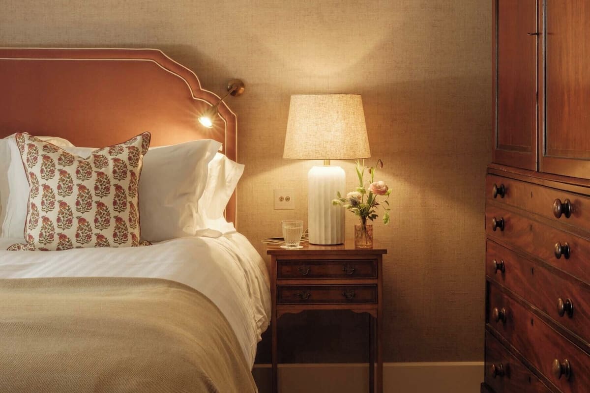 Een netjes opgemaakt bed met patroon kussens, een nachtkastje met een lamp en een vaas bloemen, en een houten dressoir.