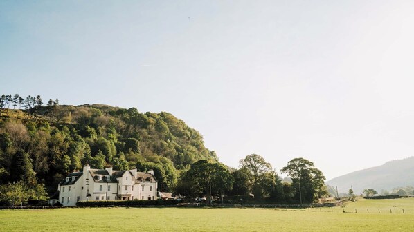 A large white building with multiple chimneys, surrounded by a green field and trees, with a hill in the background. - Fortingall (Aberfeldy)