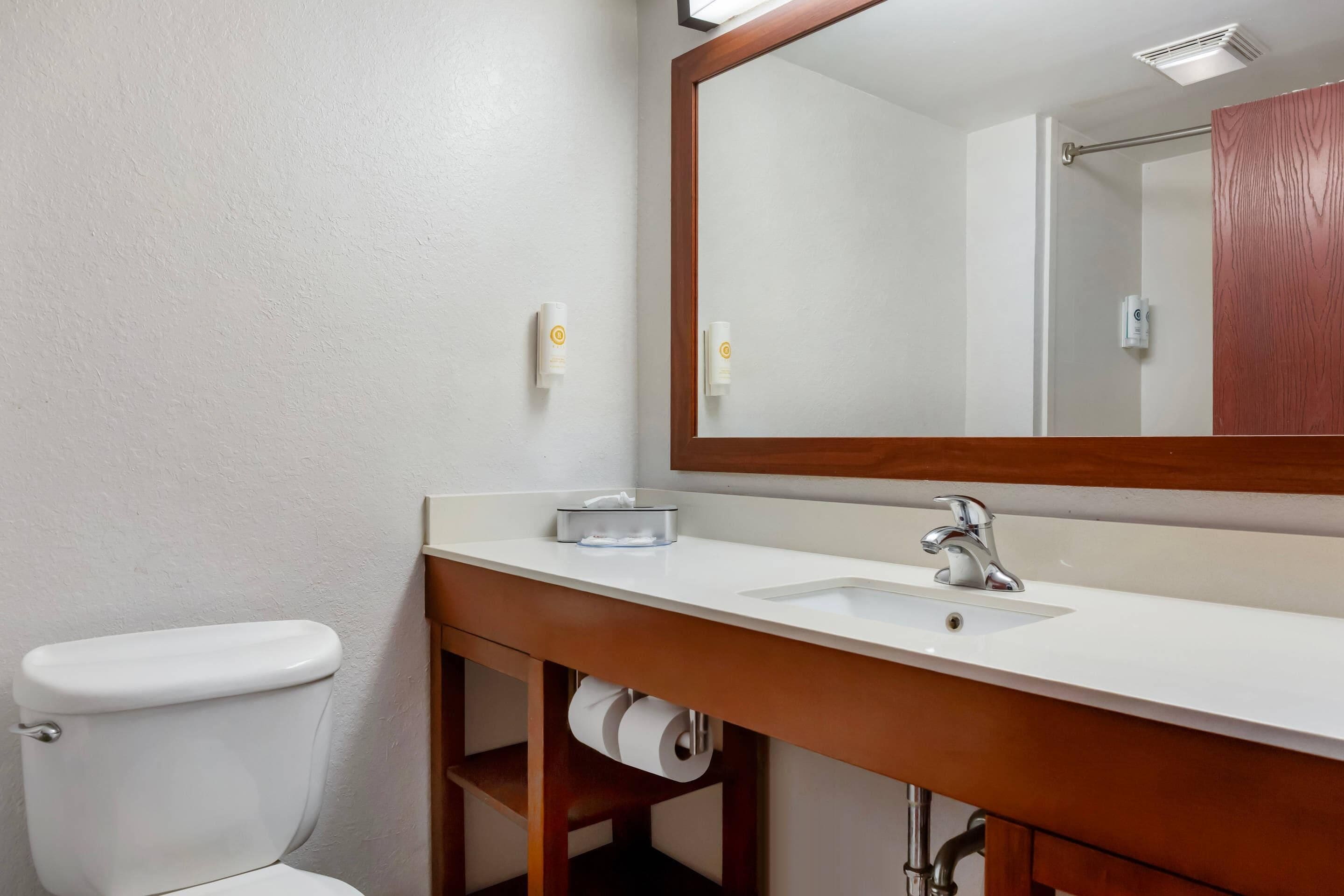 Un baño con inodoro blanco, tocador de madera con lavamanos blanco y espejo arriba, y dispensador de toallas en la pared.
