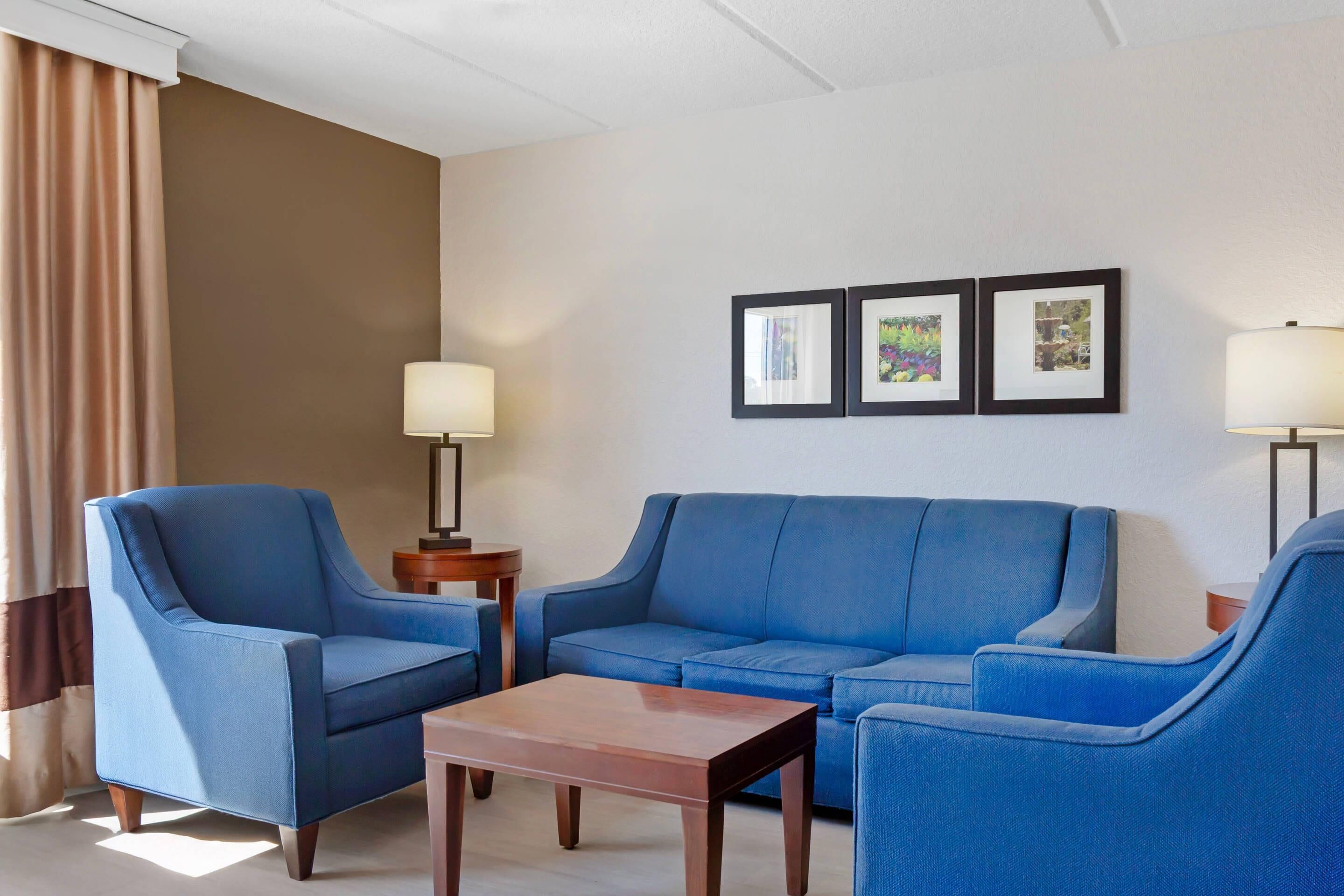 A hotel room with a blue armchair, a blue sofa, a wooden coffee table, and two floor lamps. There are three framed pictures on the wall.